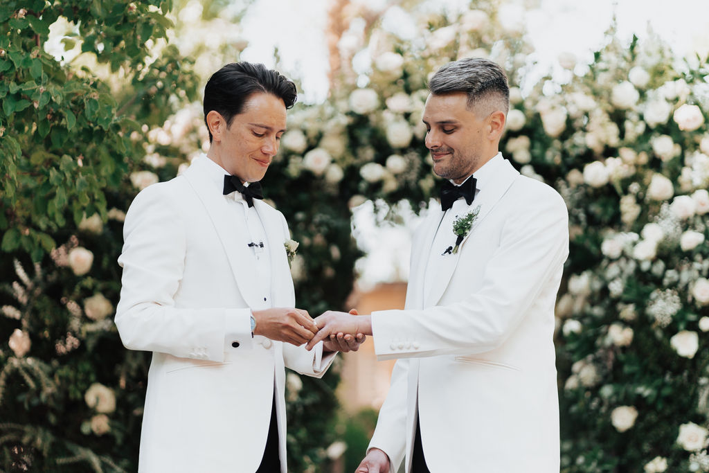 Isaac (left) putting a copper ring on Joe's (right) left hand during the ceremony standing in front of a white floral arch.
